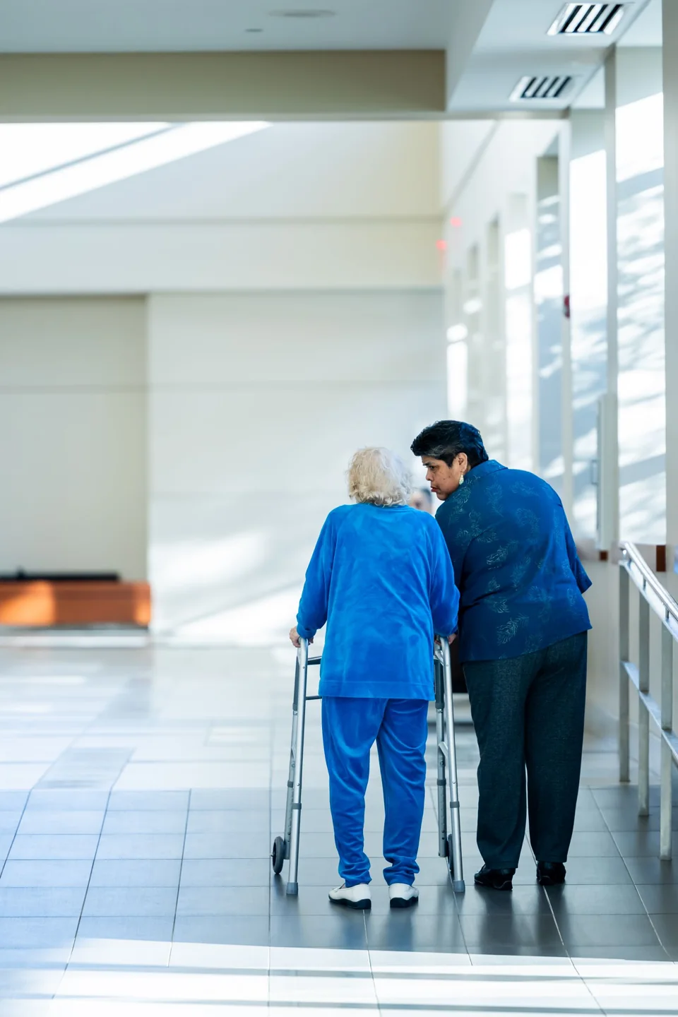 An older woman using a walker with an attendant beside her in a bright facility corridor